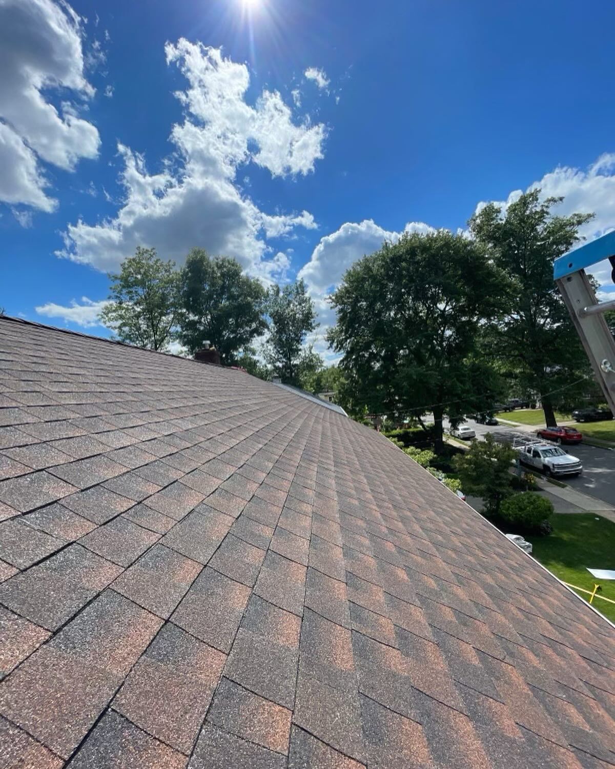 A shingled rooftop on a sunny day with trees in the background, a bright blue sky, and fluffy white clouds.