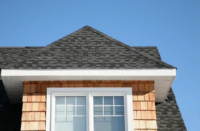 Close-up of a house corner featuring a dark-shingled roof, white trim, and wood siding around a window, against a clear blue sky.
