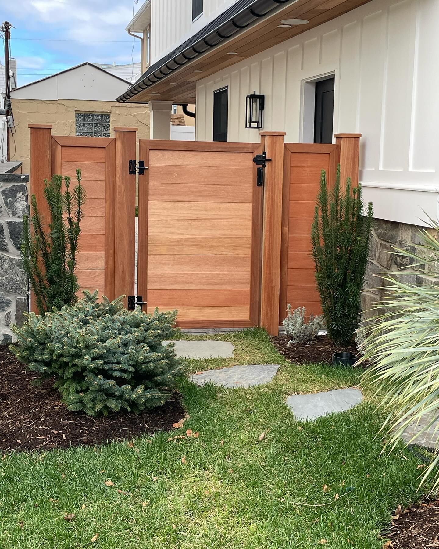 Wooden gate in a yard with a path leading to it, surrounded by greenery, and a white house in the background.