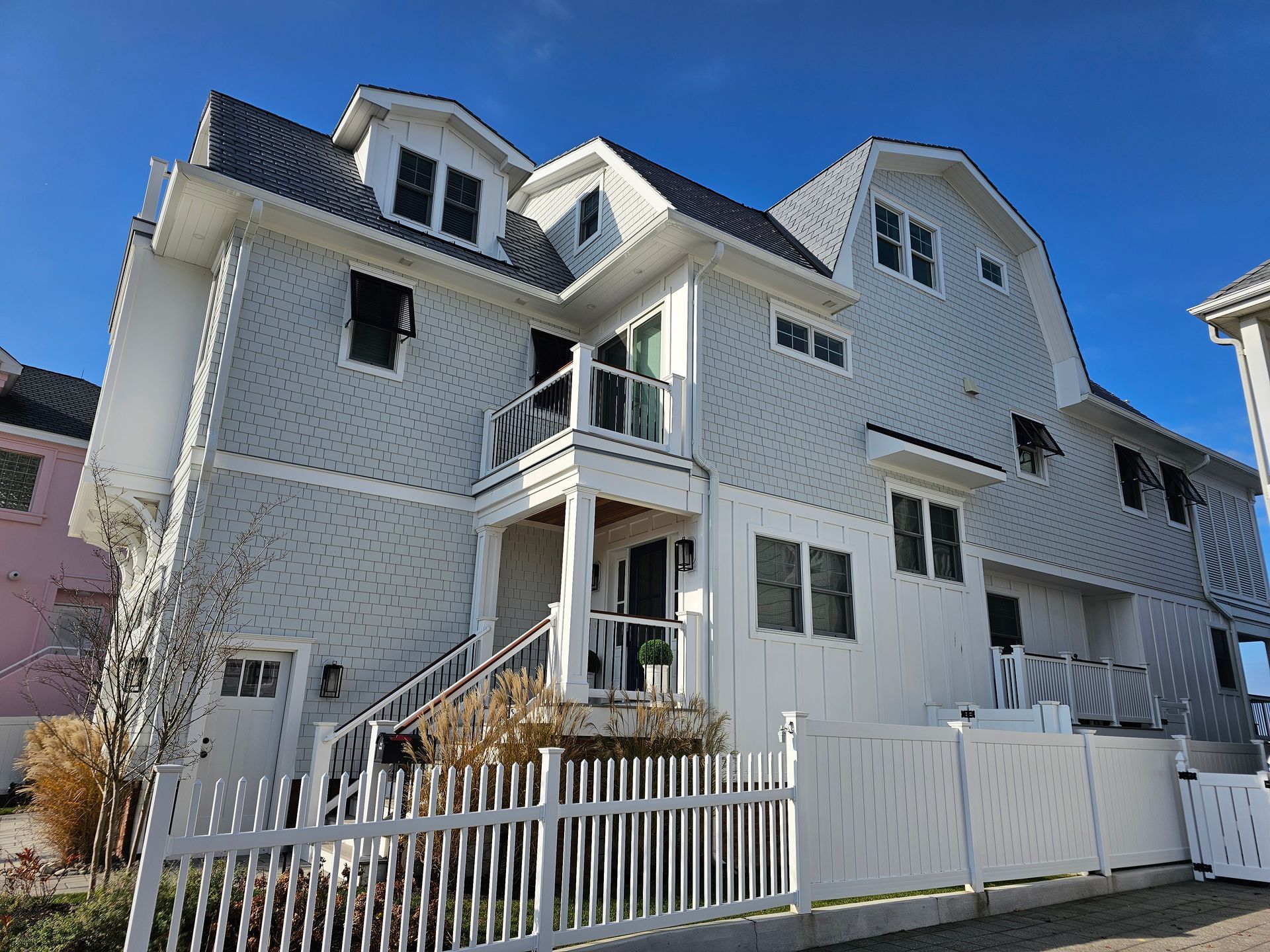 A three-story, light-grey house with a white picket fence in front of a bright blue sky.