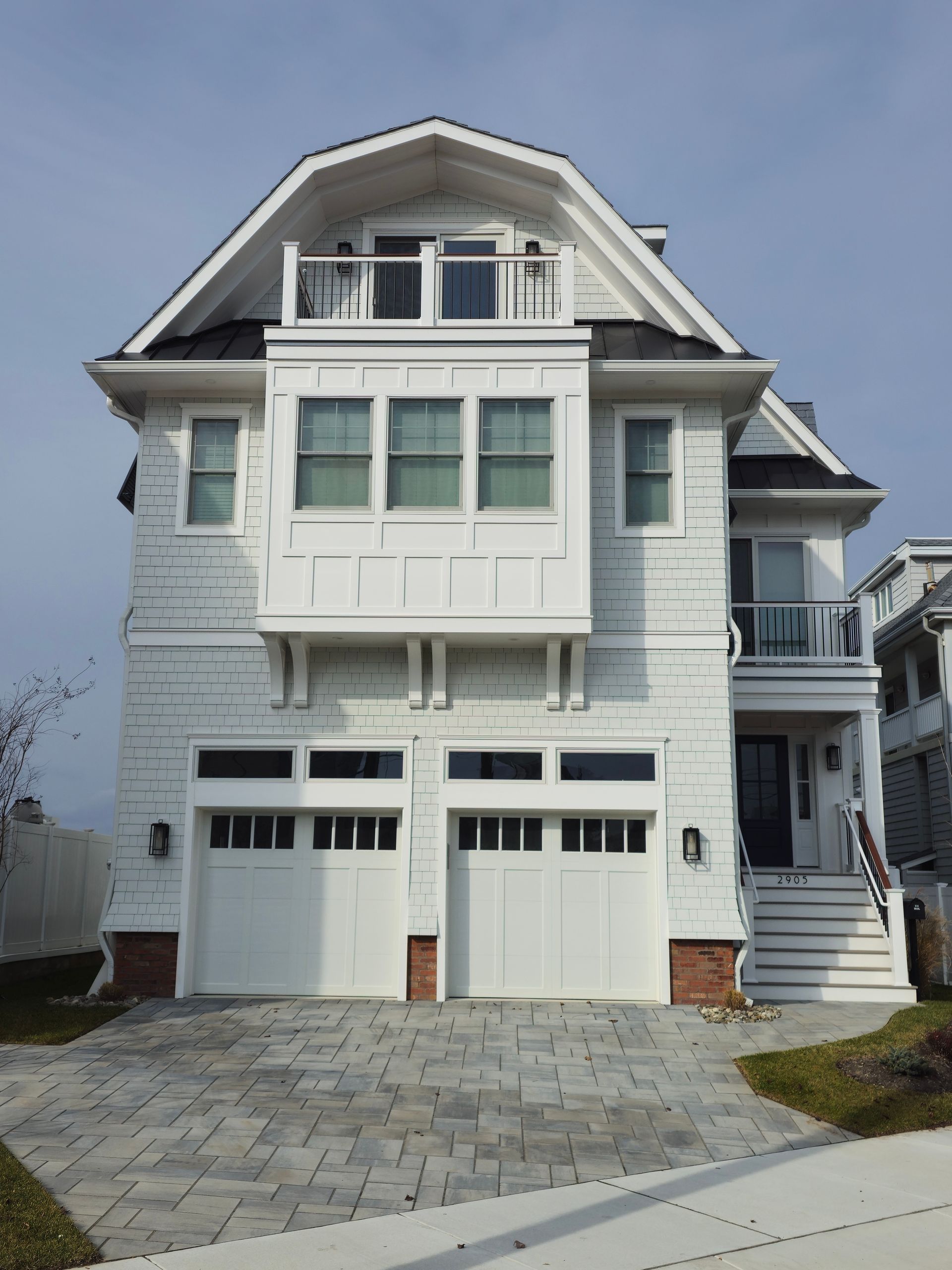 White beach house with a two-car garage, a balcony, and a gabled roof against a hazy sky. Brick accents add warmth to the facade.