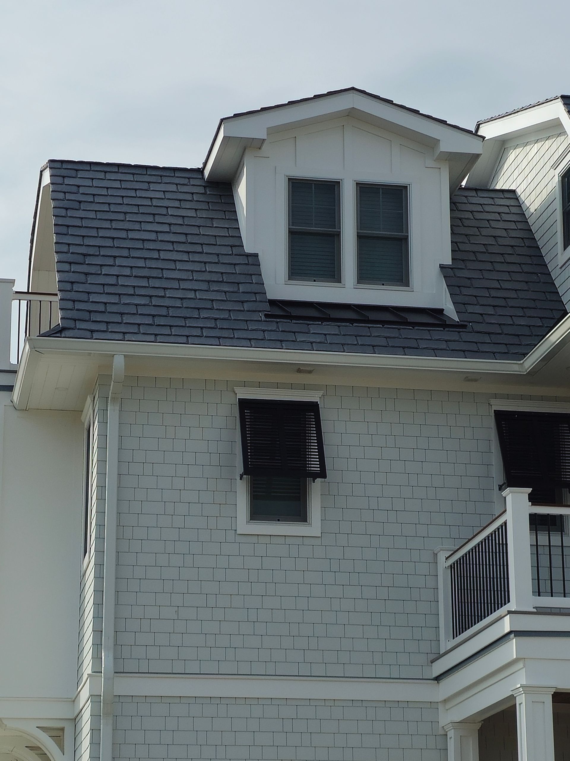 Two-story house with white shingles, dormer windows, and dark roof. One window has open black shutters.