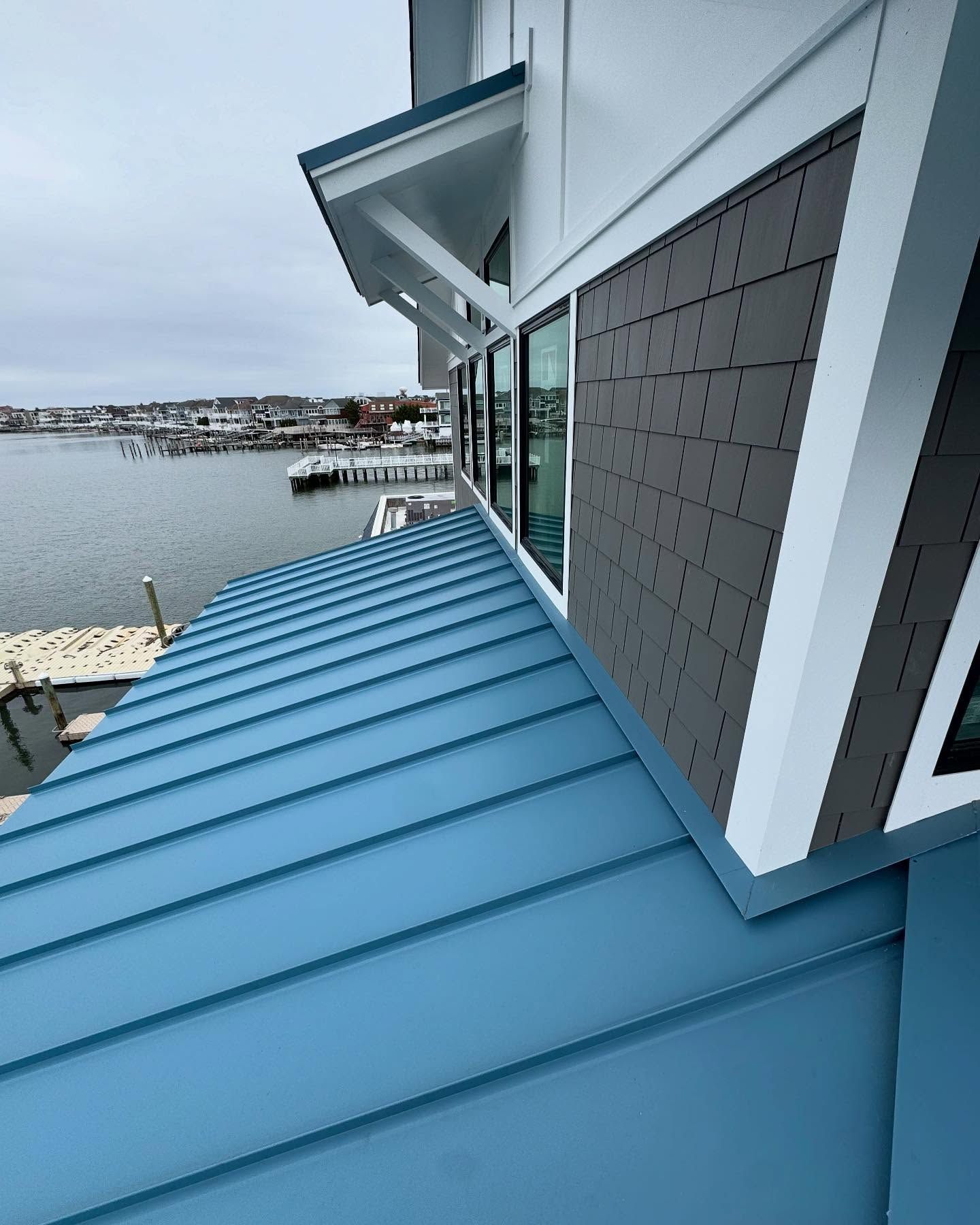 Blue metal roof of a building with blue and gray siding, overlooking a harbor with boats on an overcast day.