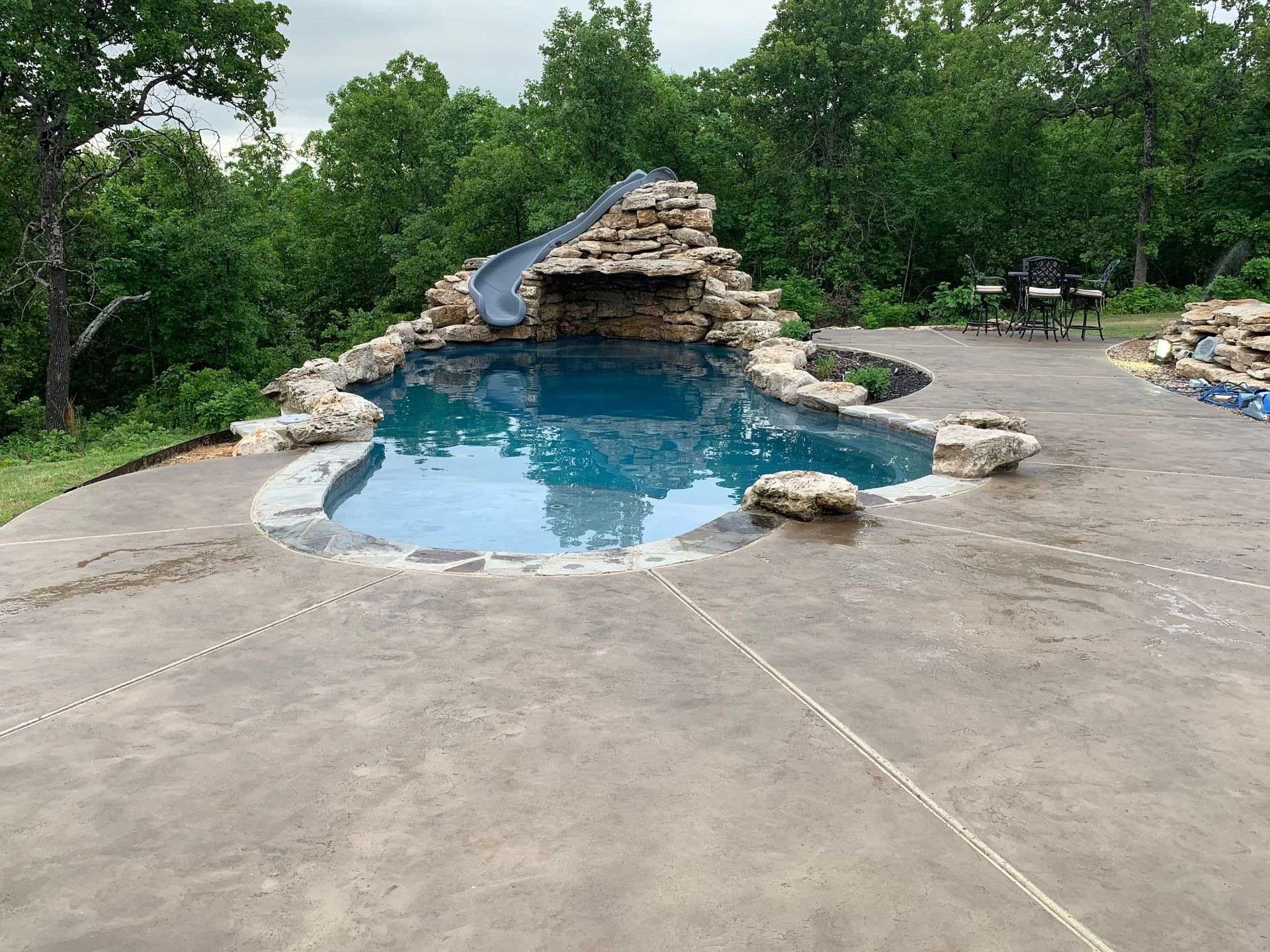 Swimming pool with rock waterfall and slide, surrounded by a concrete patio and trees.
