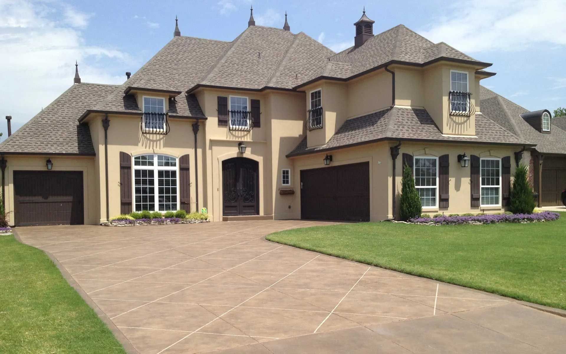 Beige two-story house with brown accents, shutters, and a driveway. Lush green lawn and gray roof.