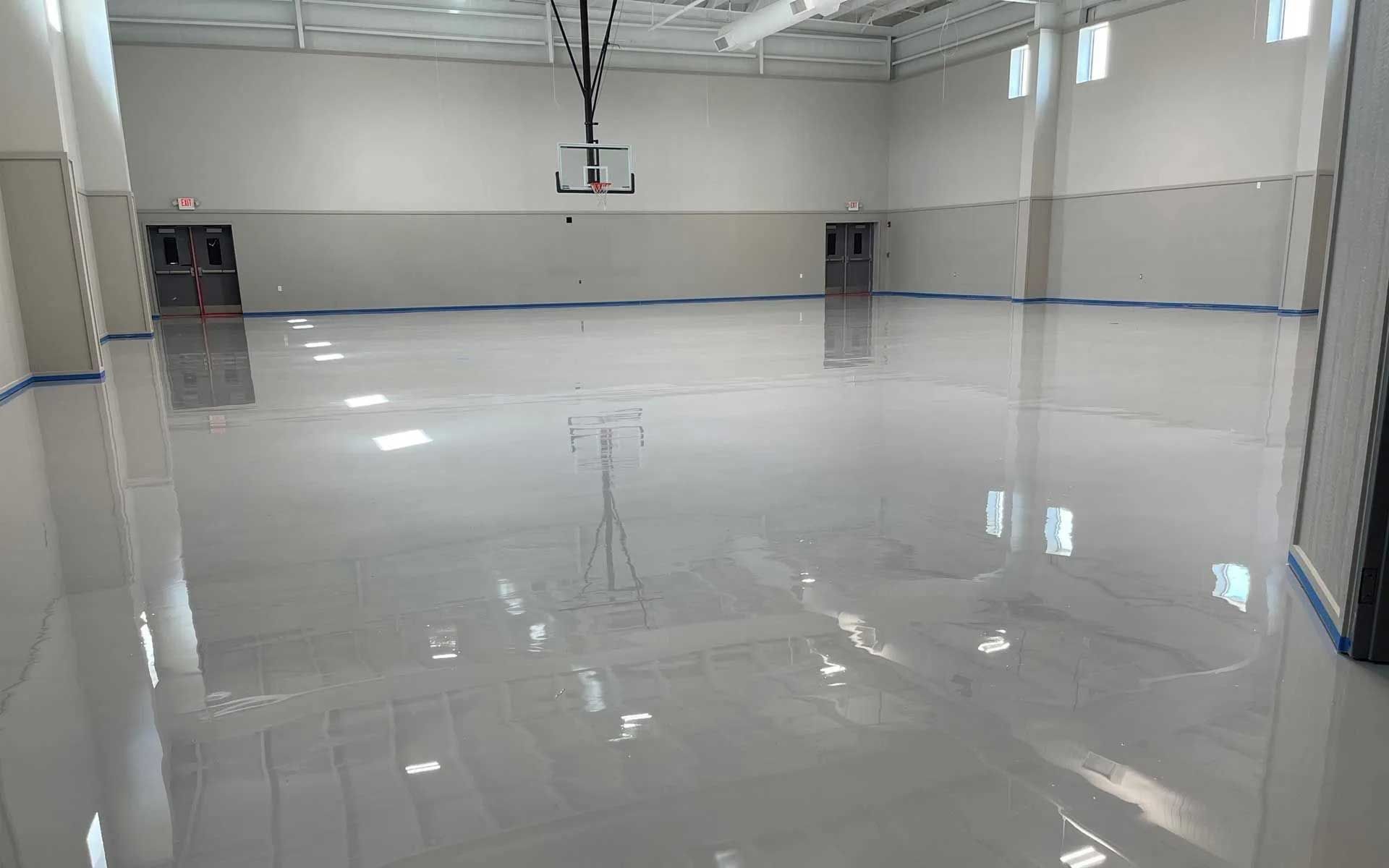 Empty indoor gymnasium with glossy gray floor, basketball hoop, and closed doors.