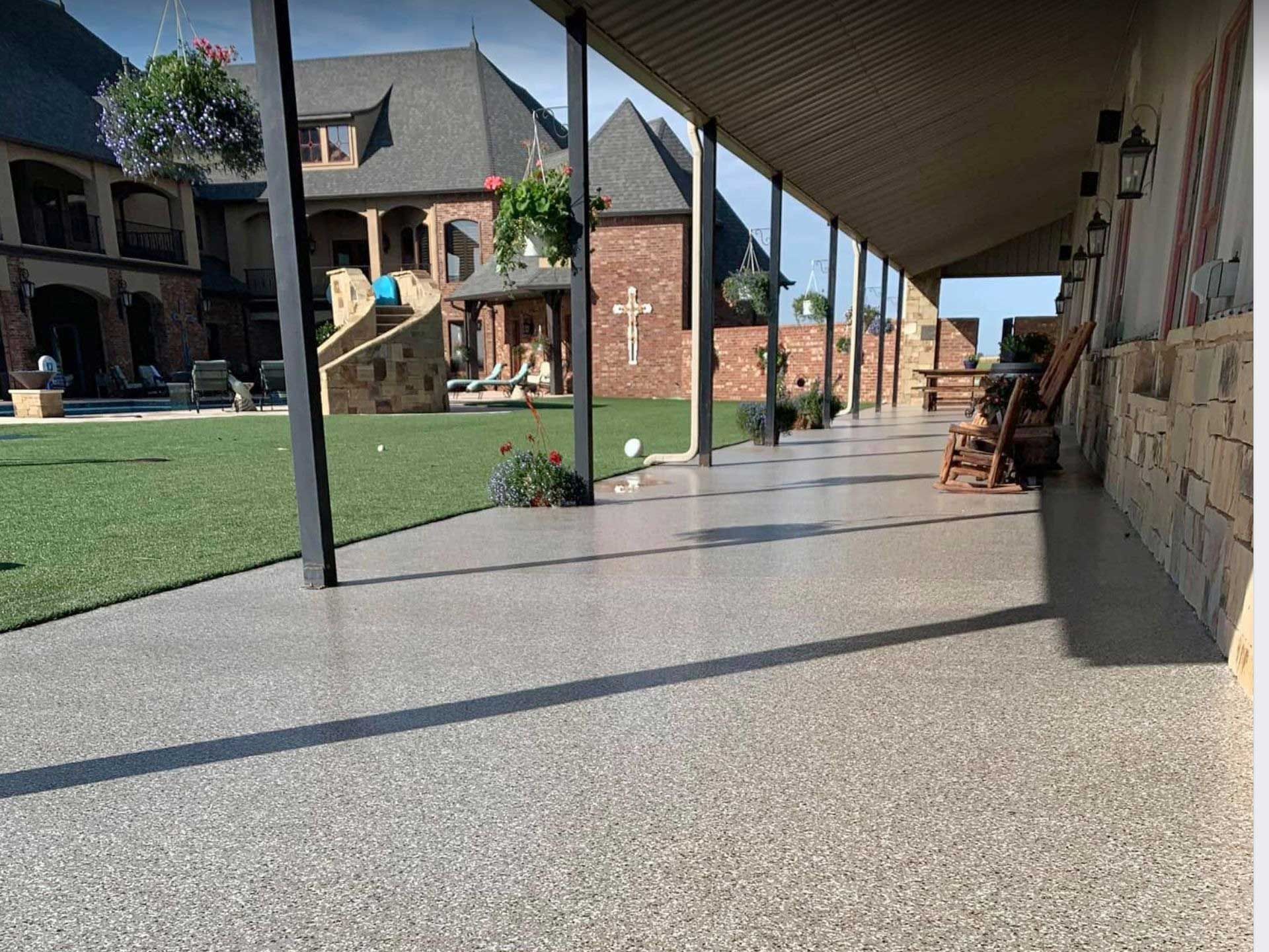 Covered patio with speckled gray floor and view of a large house with lawn and hanging flowers.