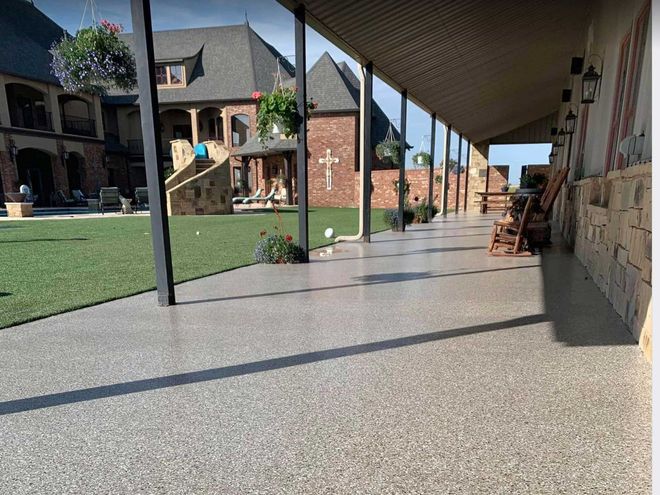 Covered patio with speckled gray floor and view of a large house with lawn and hanging flowers.