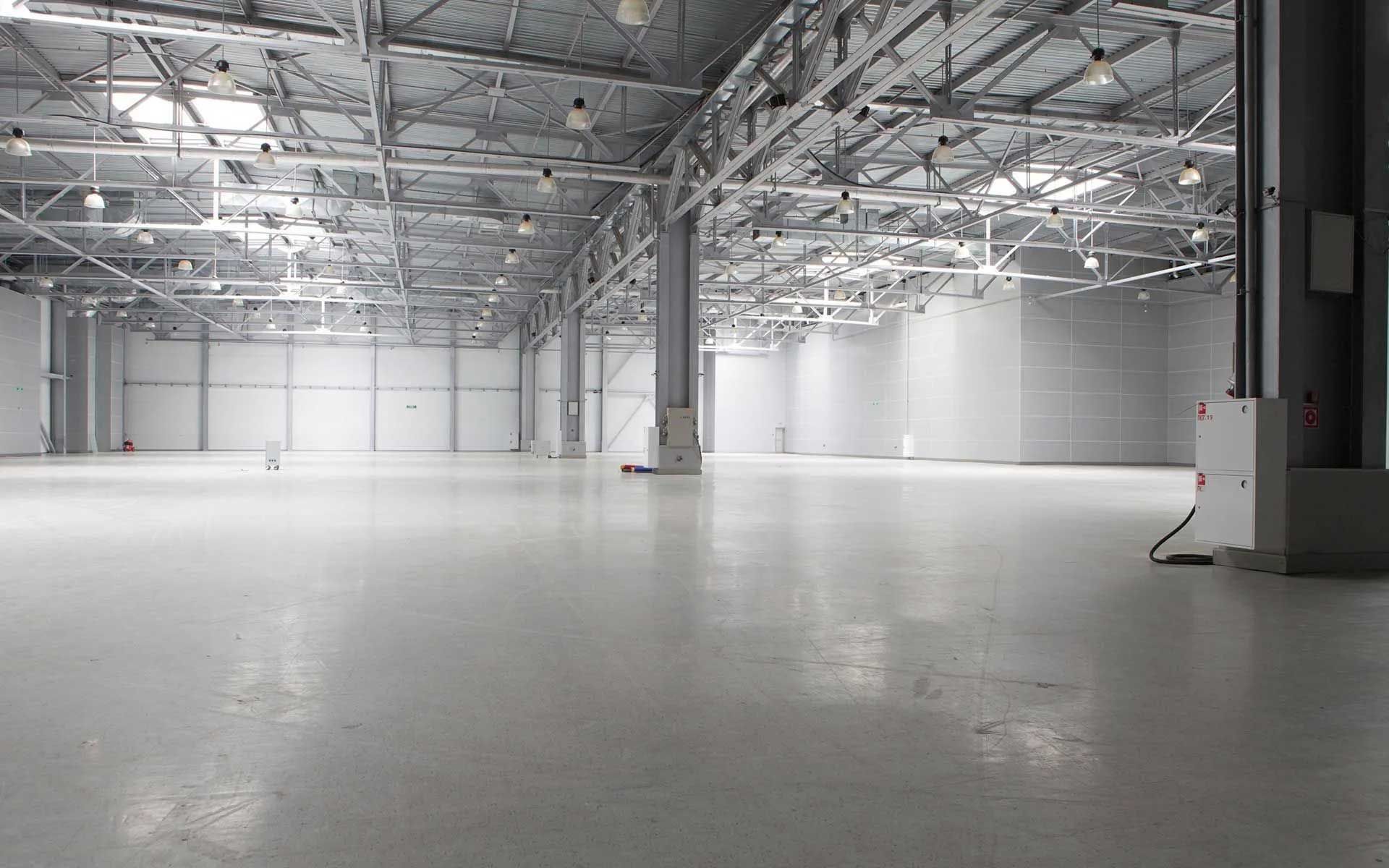 Large, empty warehouse interior with bright white floor and walls, metal beams, and ceiling lights.