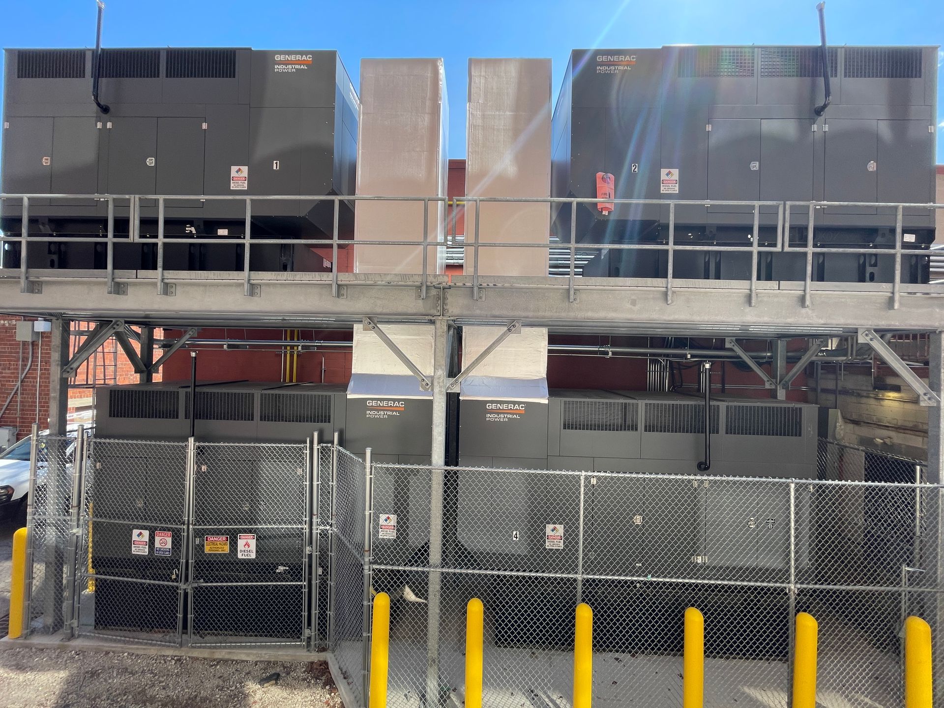 Industrial HVAC units on a rooftop behind a chain-link fence and yellow bollards