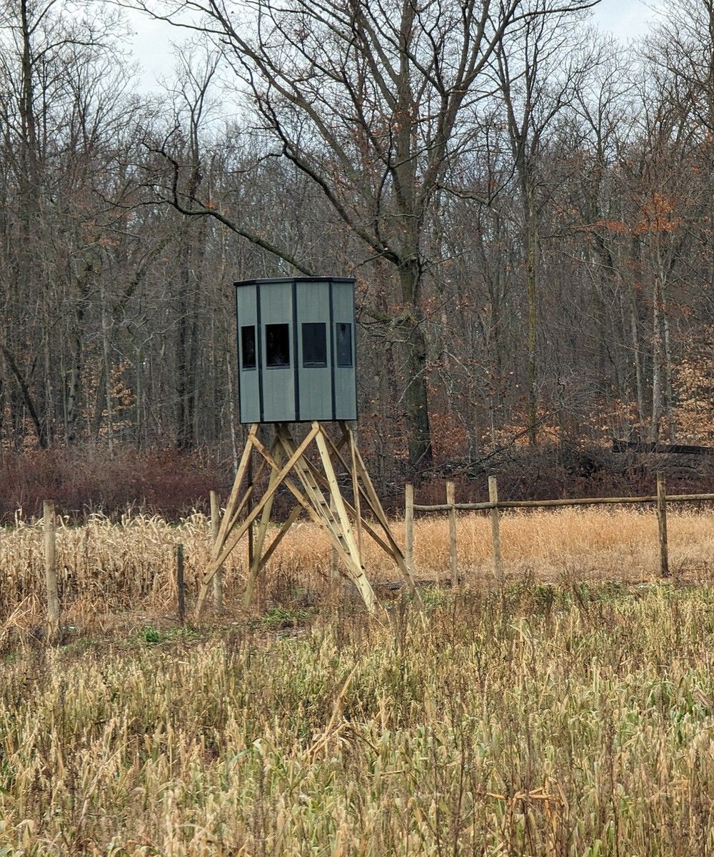 Hunting blind on wooden legs in a field with a wooded backdrop.