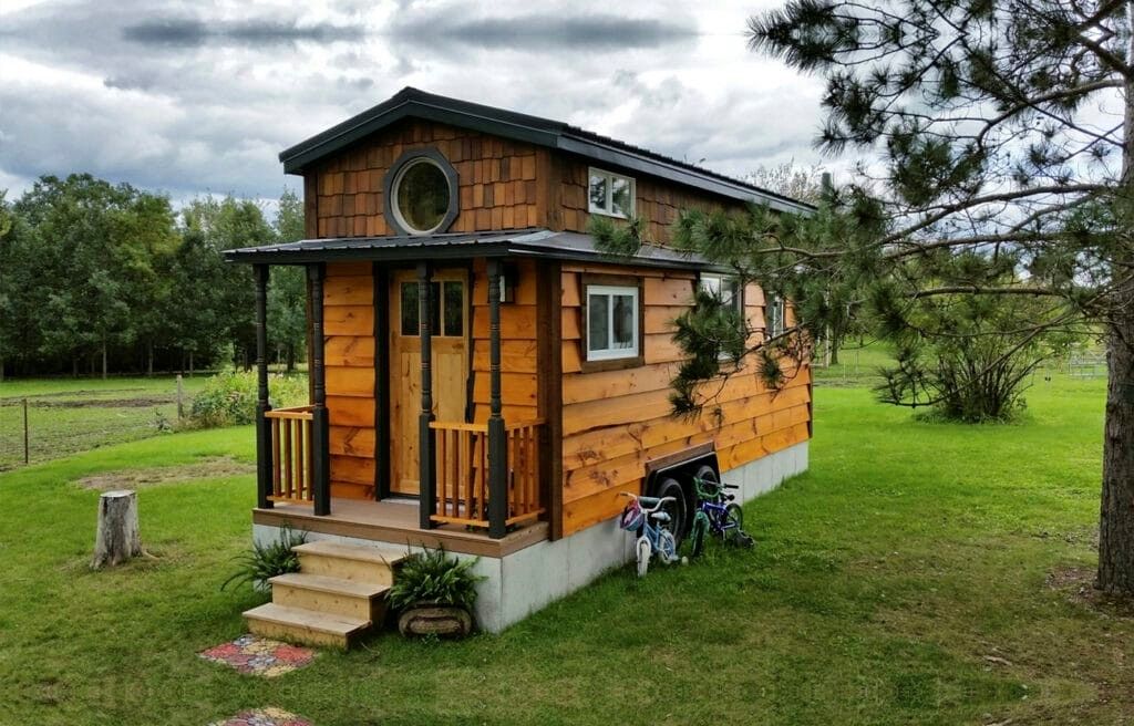 Tiny wooden house with porch, round window, surrounded by green grass and trees. Two bikes below.