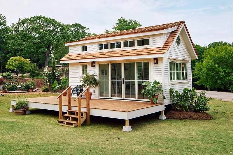 White tiny house with a brown roof and deck on a grassy lawn, trees in the background.