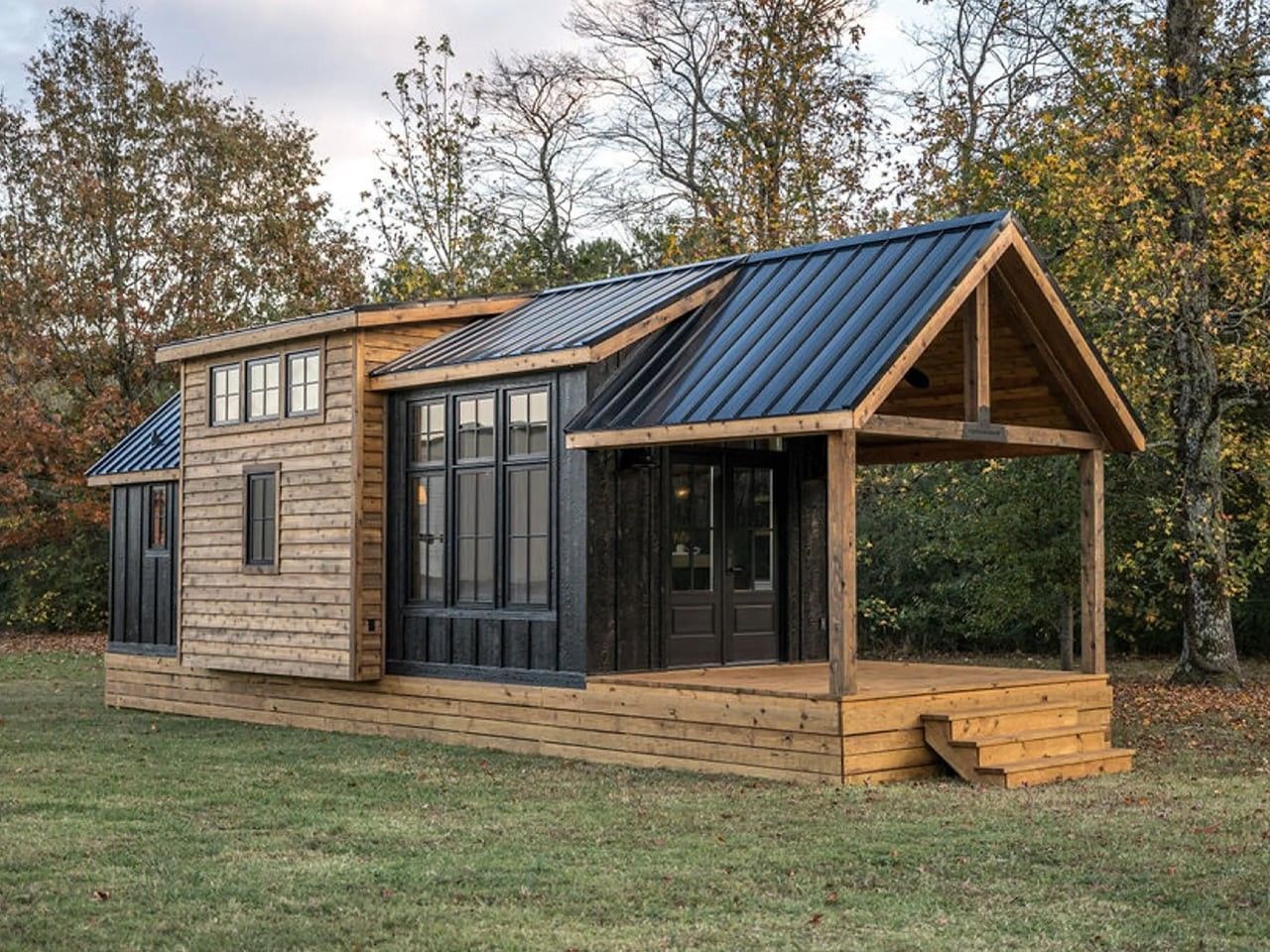 Tiny cabin with black metal roof, porch, and wooden siding, set in a grassy yard with trees.