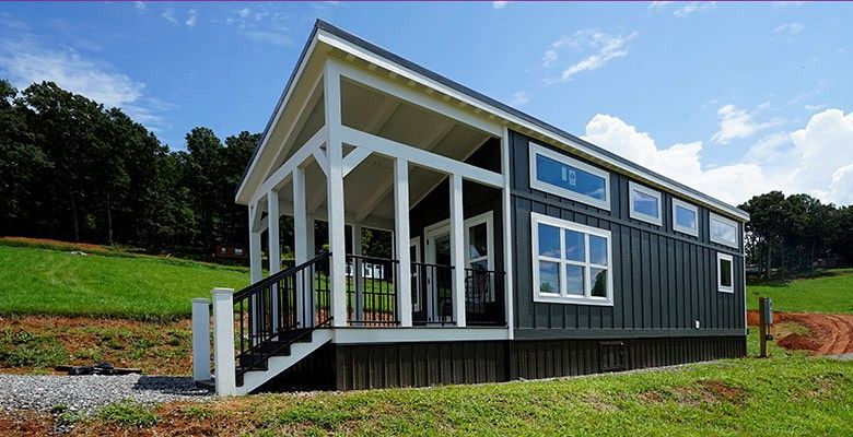 Tiny dark gray cabin with white trim, porch, and small windows on a grassy hill.