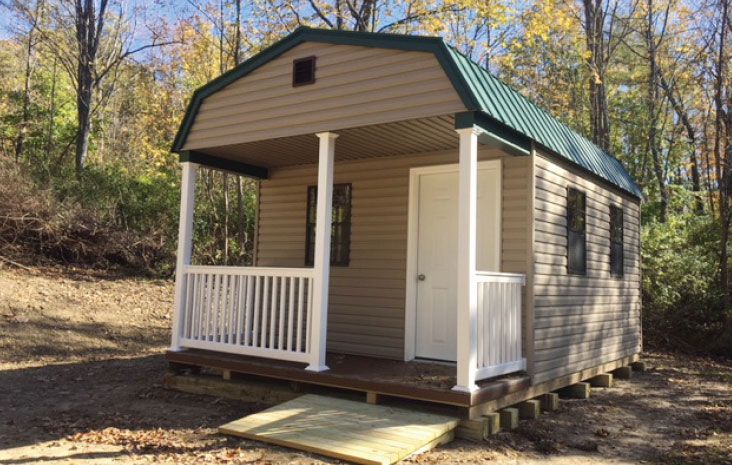 Tan shed with green roof and small porch, set in wooded area.