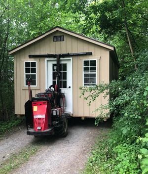 Tan shed being moved on a gravel road by a red tractor in a wooded area.