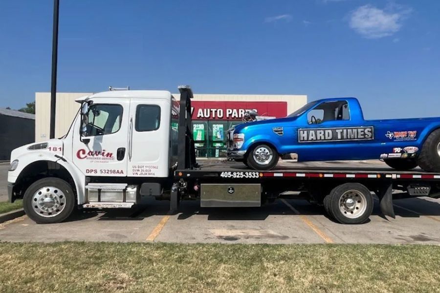 A tow truck with a blue truck on the back is parked in a parking lot.