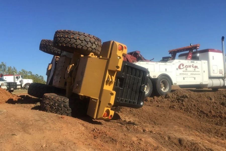 A tow truck is towing a tractor that has fallen over in the dirt.
