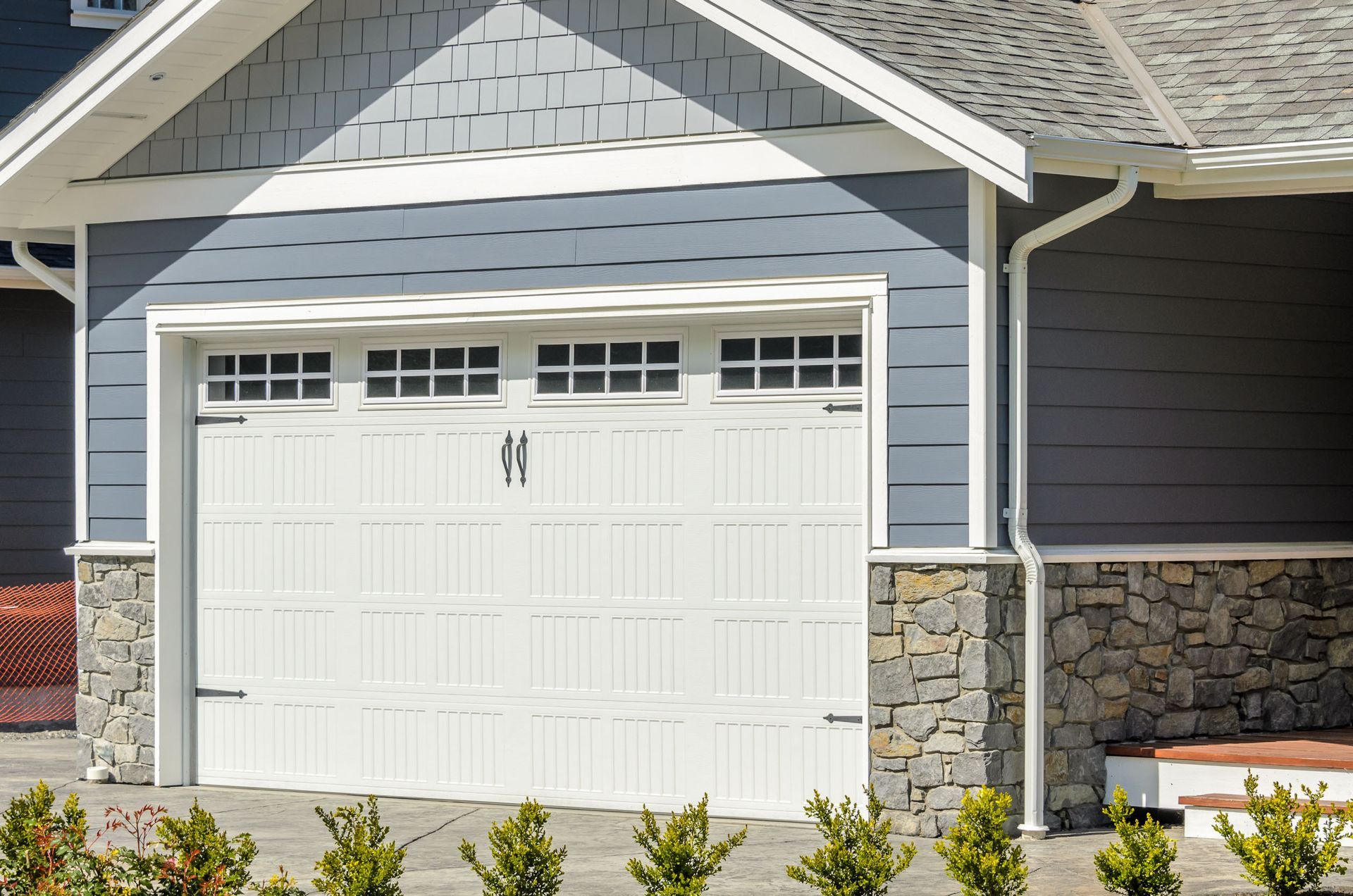 White double garage door on a gray house with stone trim and small shrubs in front.