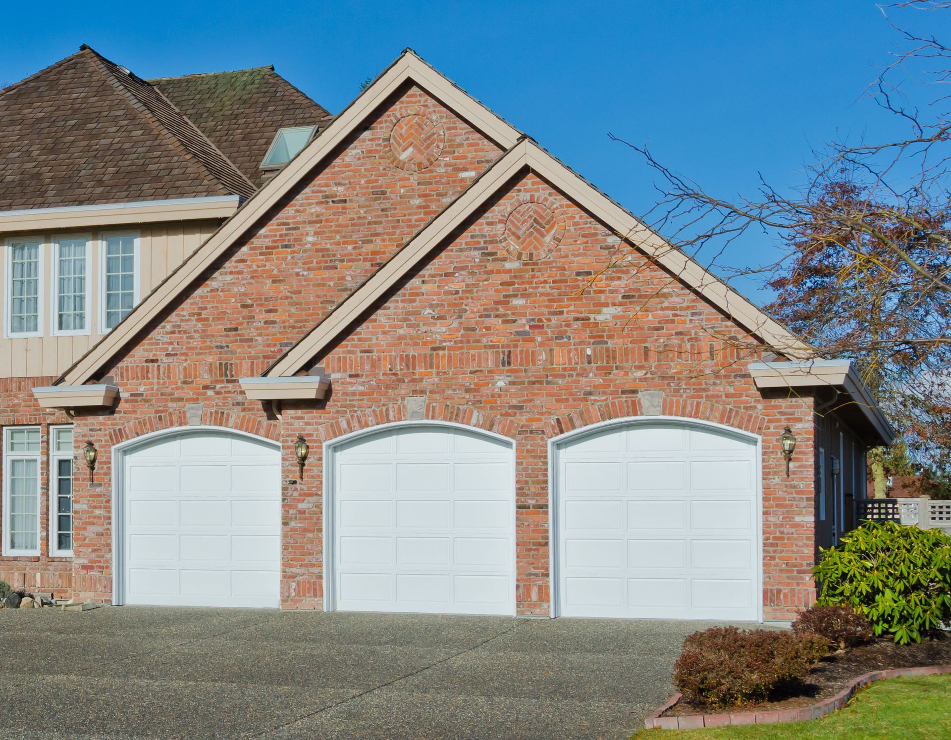 Brick suburban house with three white garage doors and a gravel driveway under a clear blue sky