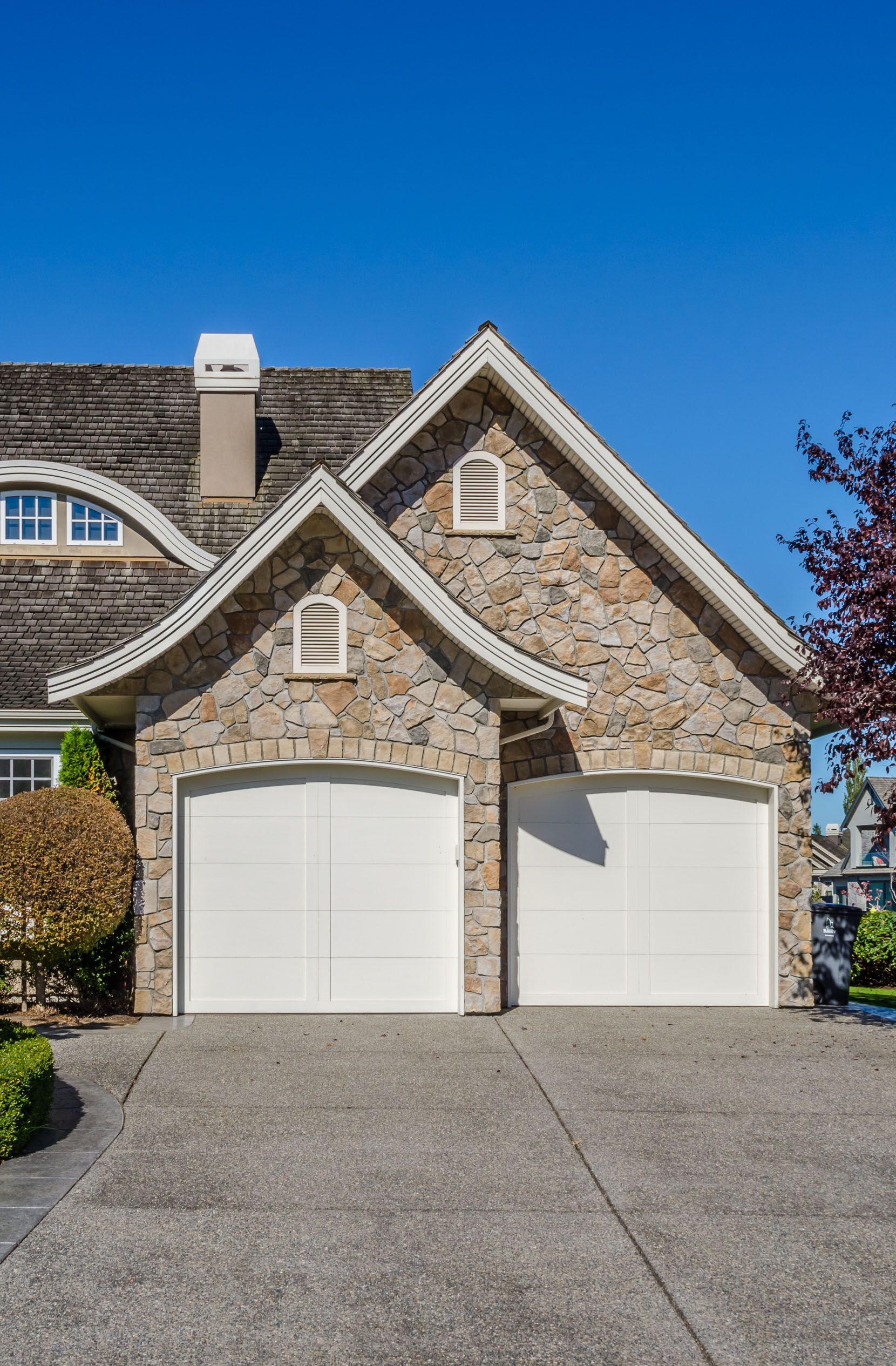 Stone suburban house with two white garage doors and a paved driveway under a clear blue sky
