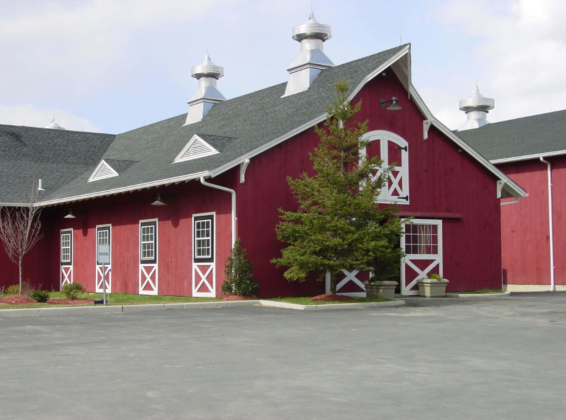 Red barn-style building with white trim and a gray roof on a paved lot, under a cloudy sky