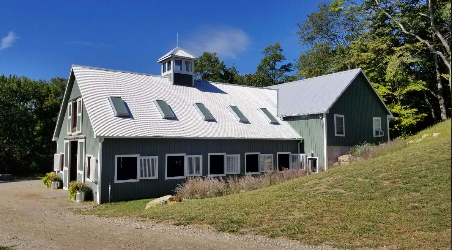 White farmhouse with black trim and dormer windows on a grassy hillside under a blue sky