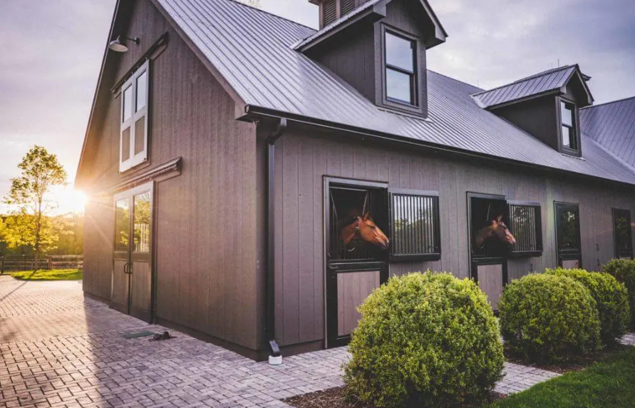 Modern gray farmhouse with lit windows, a paved walkway, and trimmed bushes at sunset