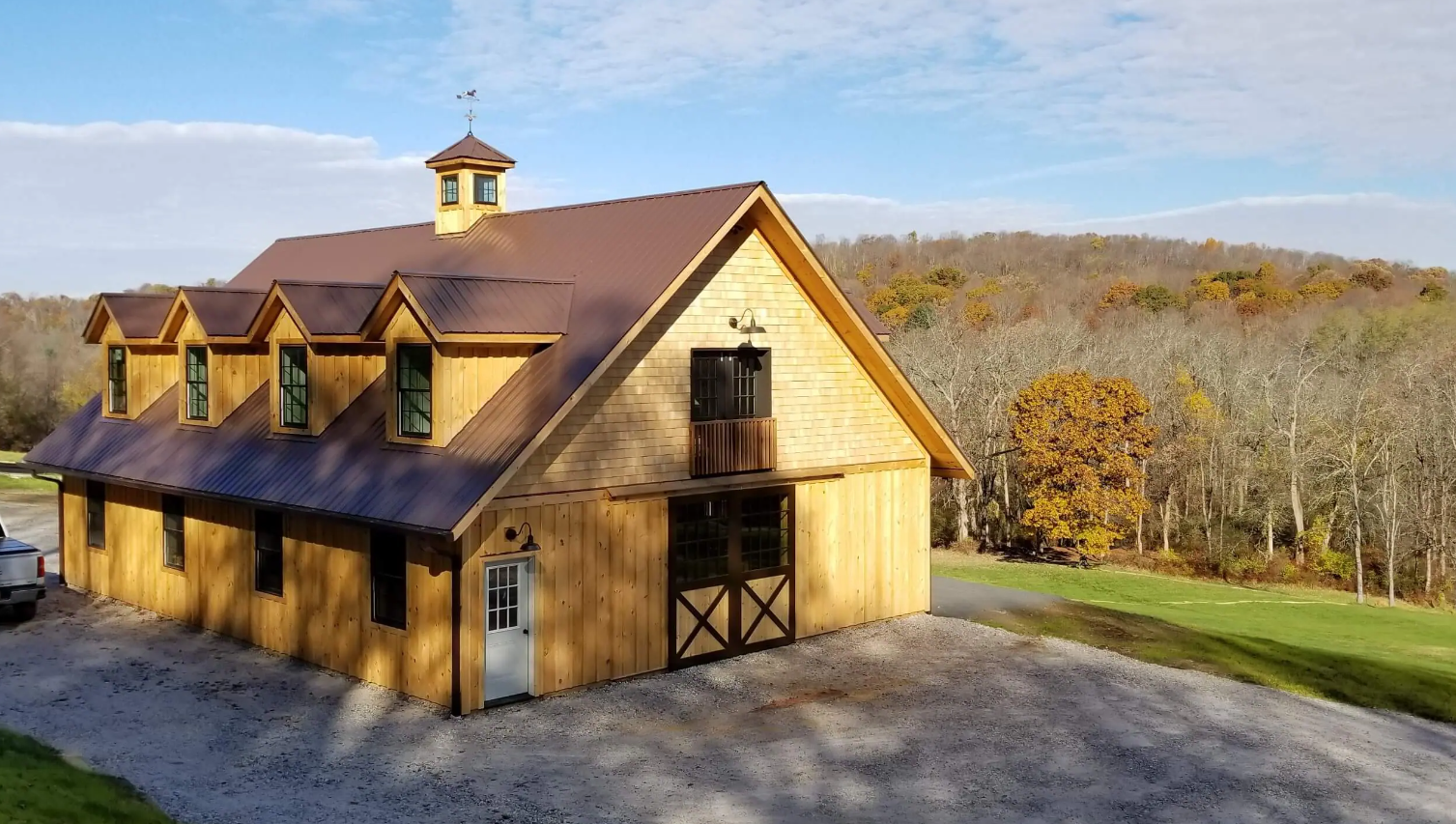 Yellow barn with brown roof on a gravel lot, set against autumn trees and a hillside.