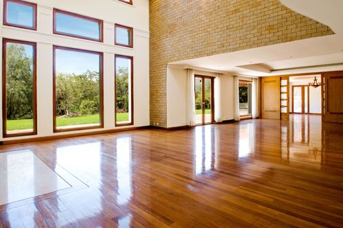Empty sunlit living room with polished hardwood floors, large windows, and stone-accent walls