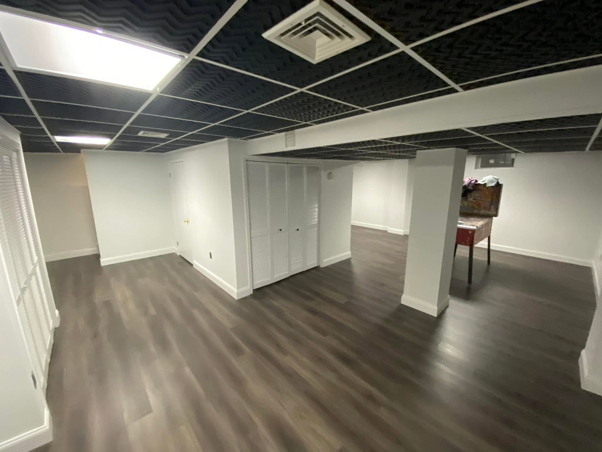 Empty office space with white walls, dark ceiling grid, and wood-look flooring.