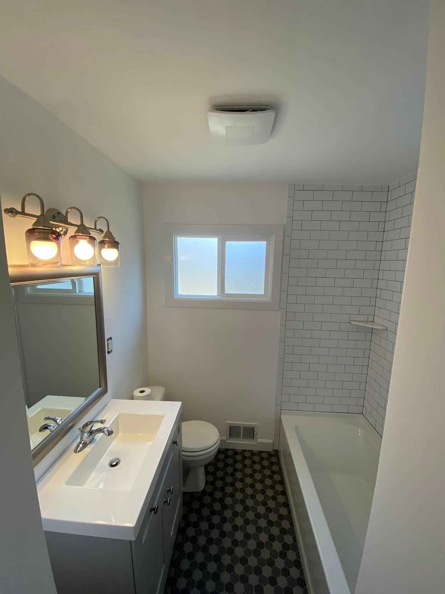 Bright bathroom with vanity, toilet, bathtub, window, and patterned tile floor.
