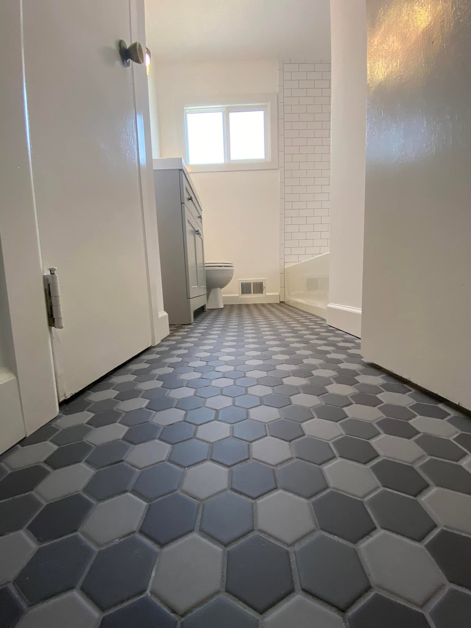 Narrow bathroom with hexagon tile floor, white walls, and a shower chair near a window.