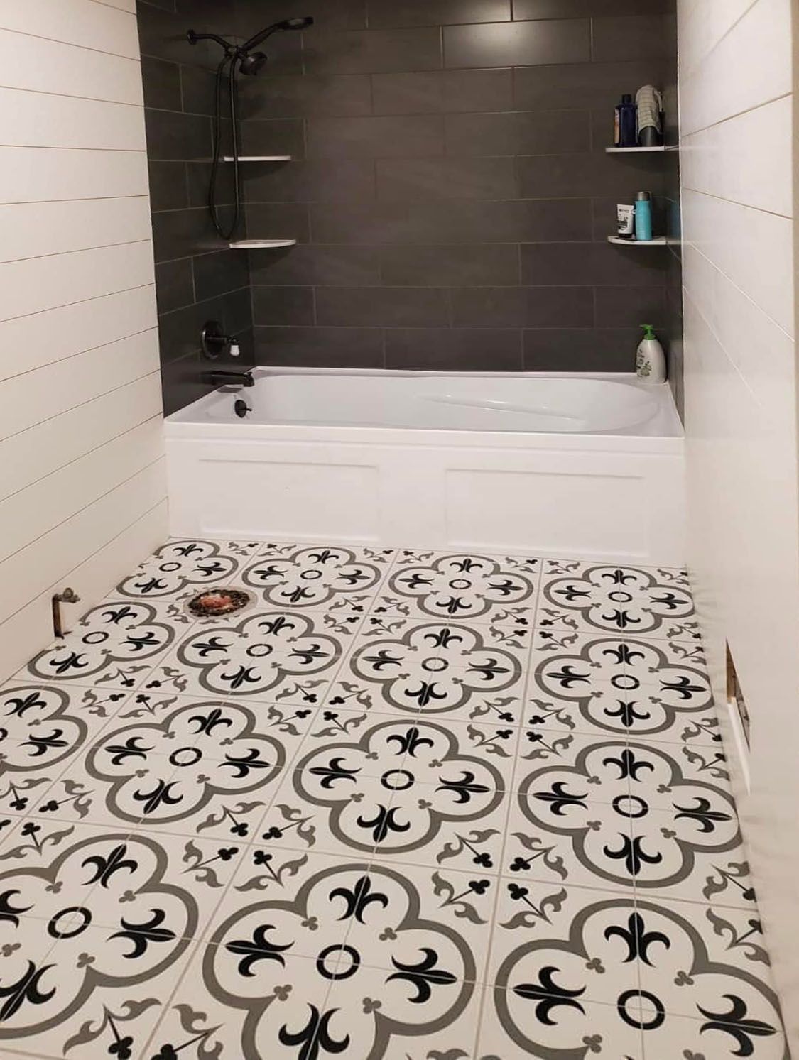 Bathroom with black-and-white patterned floor, white tub, dark shower wall, and wall shelves with toiletries.