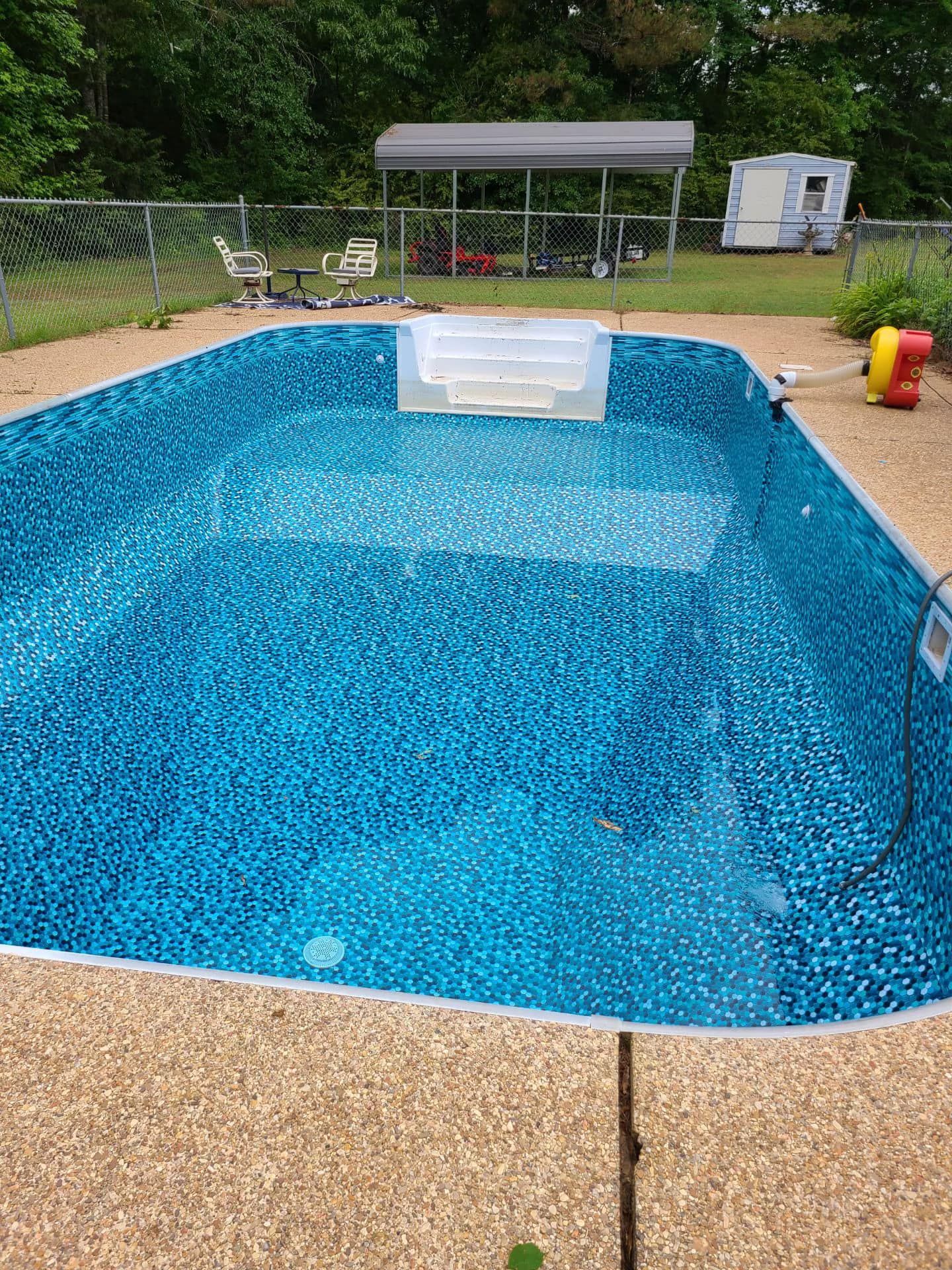Empty blue-tiled swimming pool surrounded by a concrete patio. Backyard setting with a covered shelter and toys visible.