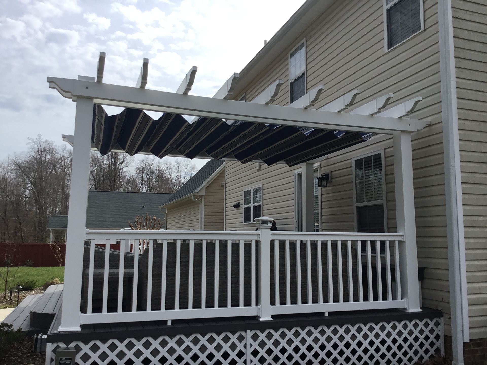 White pergola with retractable black shade over a deck attached to a beige house.