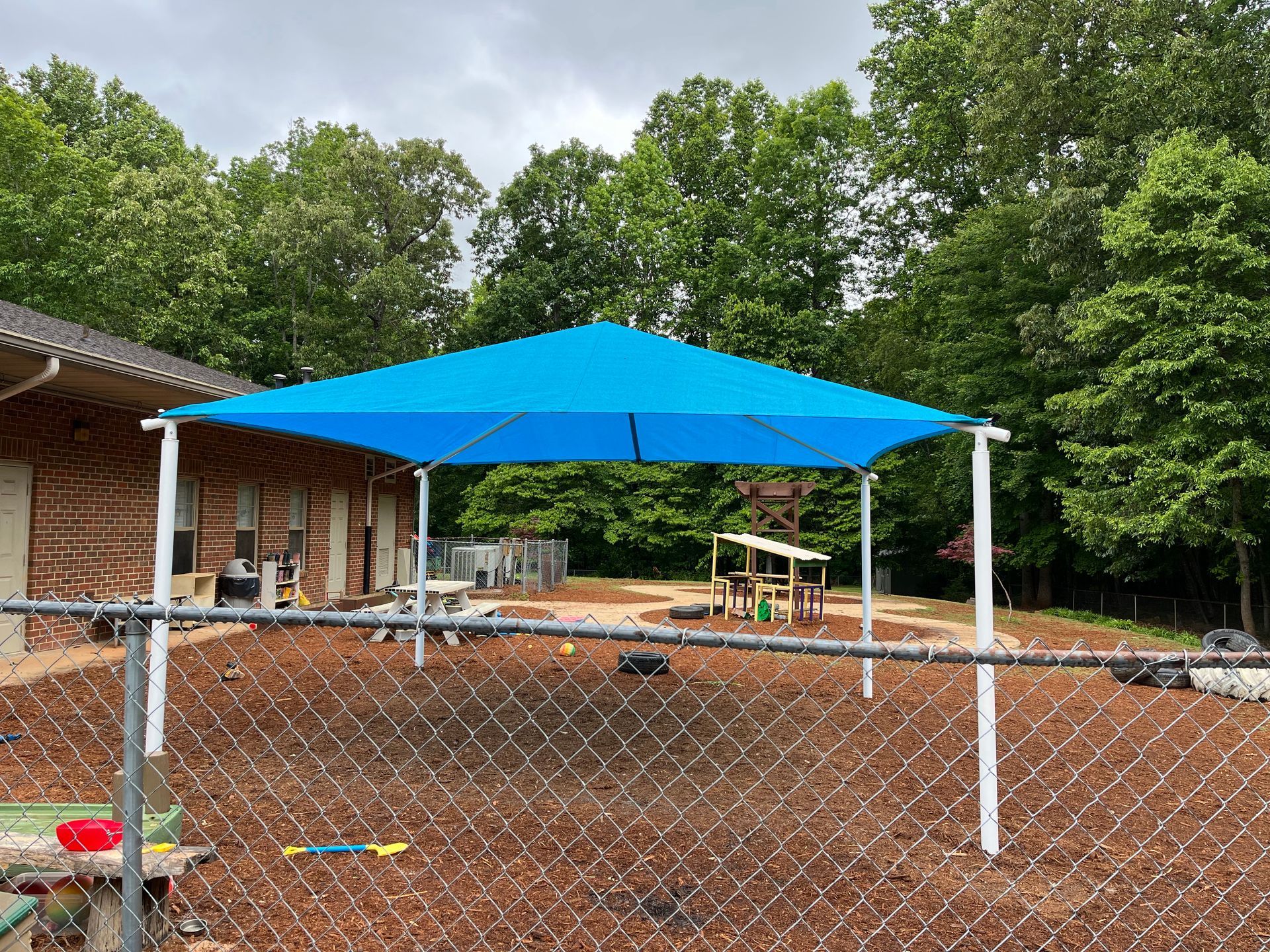 Blue canopy over a playground; white supports, fenced-in area with wood chips.
