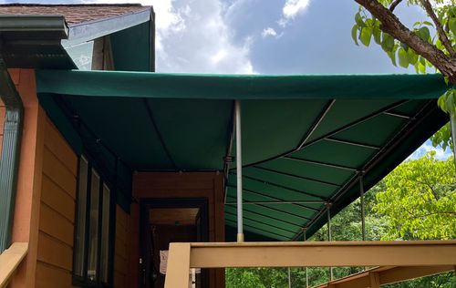 Green awning covering a wooden porch. Silver poles support the awning. Brown house and cloudy sky background.