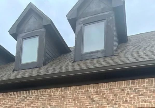 Brick house exterior with two dormer windows on a dark shingled roof and brown gutters.