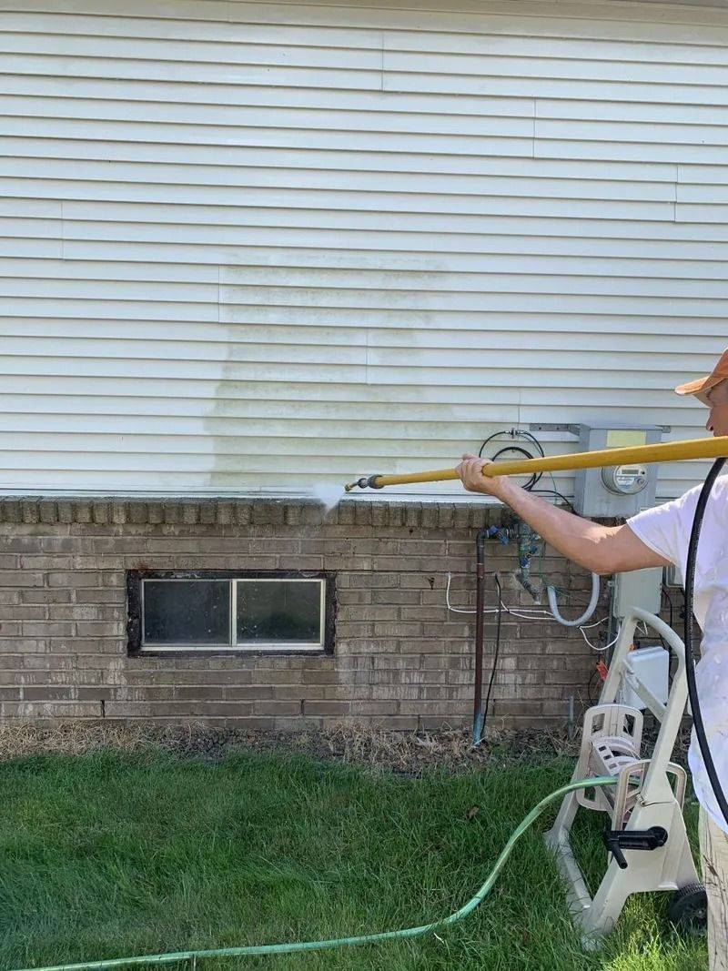 Person pressure washing a white house exterior with a brick base.