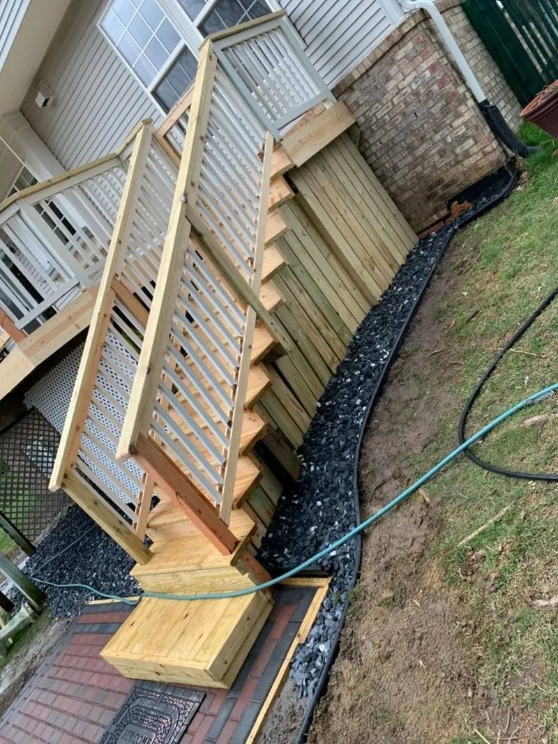 Wooden deck with stairs, brick and siding wall, dark landscaping rocks, and a green hose on the grass.