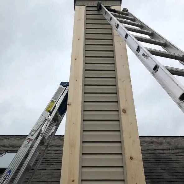 Two ladders lean against a chimney with beige siding. The sky is overcast.
