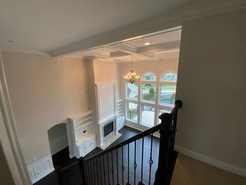 View from a staircase, overlooking a bright living room with large windows, fireplace, chandelier, and dark hardwood floors.