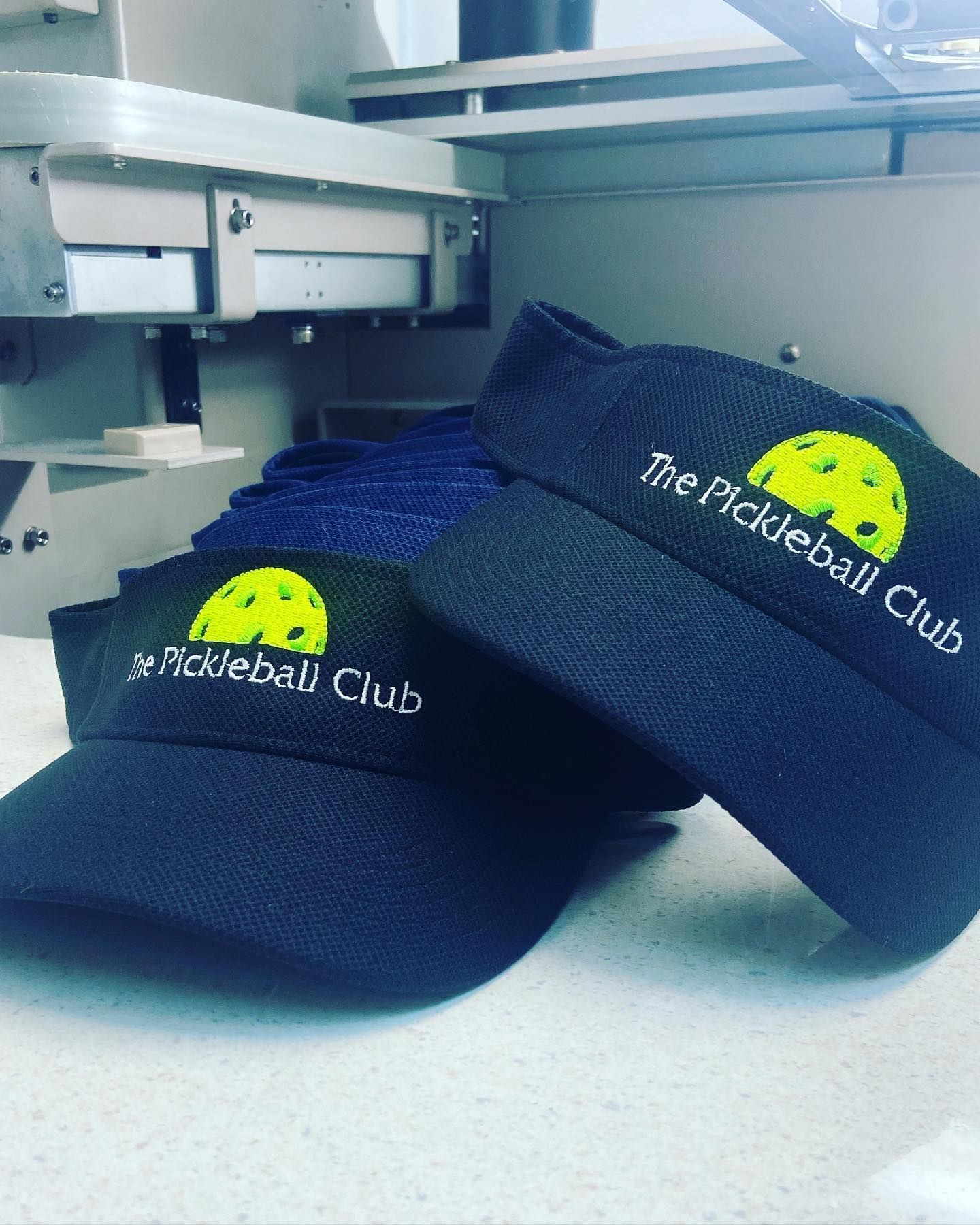Two navy visors with “The Pickleball Club” logo on a countertop