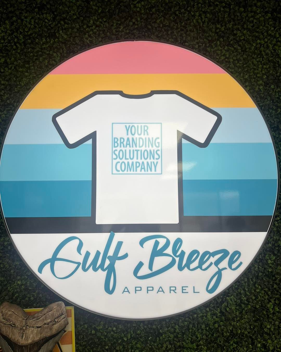 Logo for Gulf Breeze Apparel featuring a white T-shirt over a pastel sunset circle.