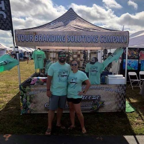 Two people posing at a branded outdoor vendor booth under a canopy tent.