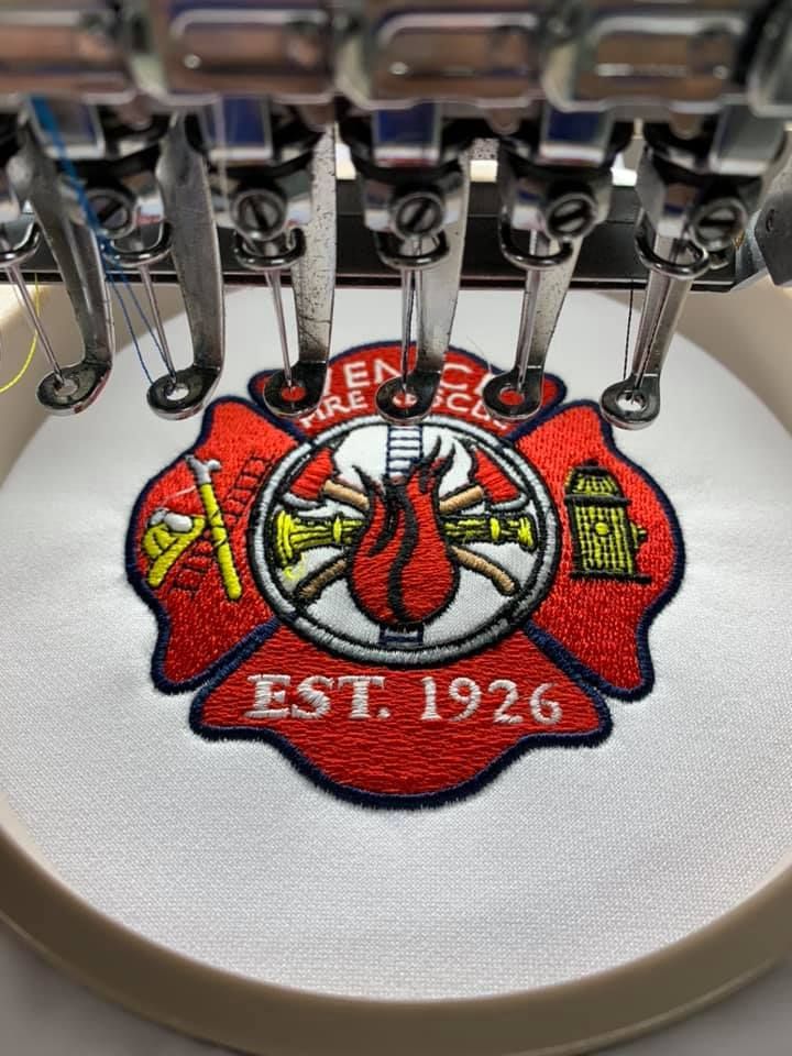 Close-up of an embroidery machine stitching a red fire department emblem with “EST. 1926” on white fabric