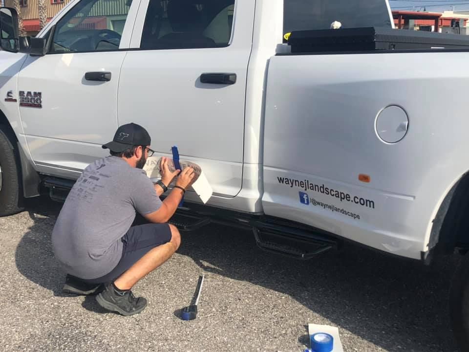 Person crouches beside a white pickup truck, using a tool near the lower side panel.