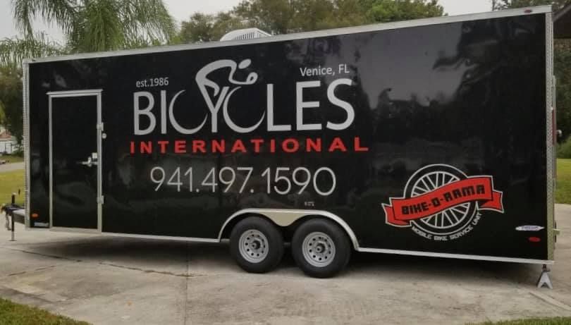 Black trailer with “Bicycles International” logo and phone number parked outdoors near trees.